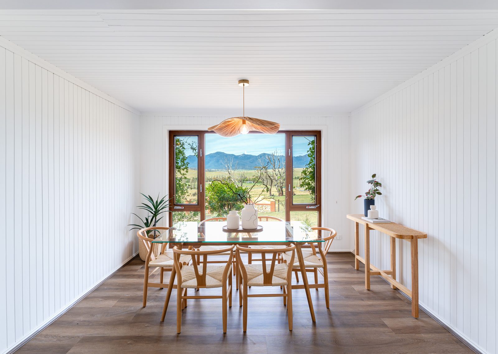 A dining room with shiplap walls, wishbone chairs around a glass table, a paper pendant overhead, and a wide window looking out to a mountain range.