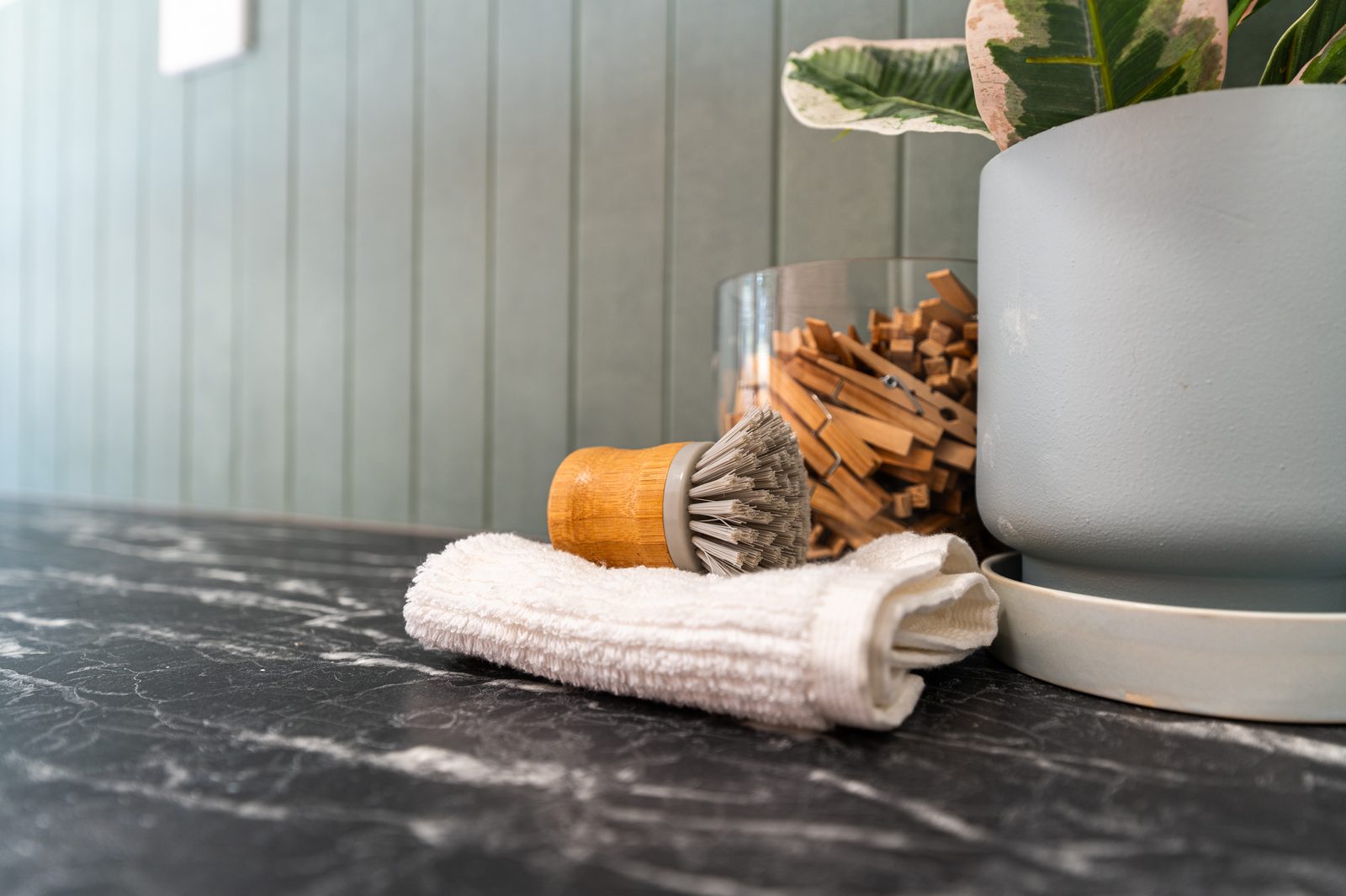A laundry bench with a jar of clothes pegs and a small plant, against a sage-green v-groove wall.