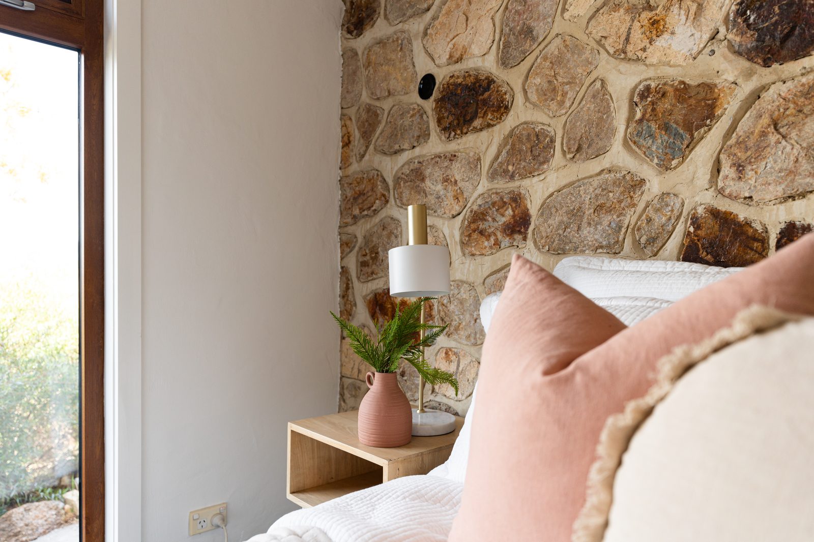 A bedroom corner with a sandstone feature wall, a terracotta jug holding a fern on a timber side table, and a soft blush cushion at the edge of the bed.
